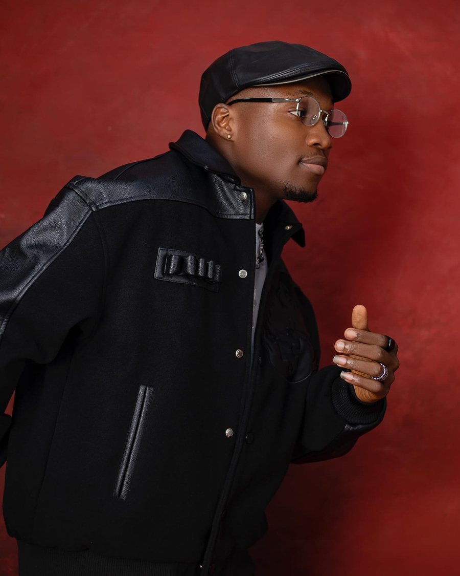 Cityman posing against a deep red background, wearing a black flat cap, round glasses and a black varsity-style jacket