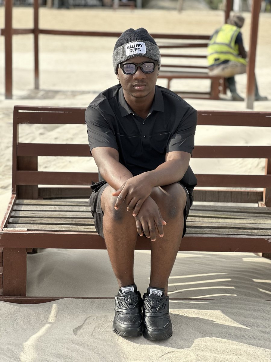 High Glory Music seated on a wooden beach lounger, wearing a grey beanie, dark sunglasses, a black polo shirt and black sneakers